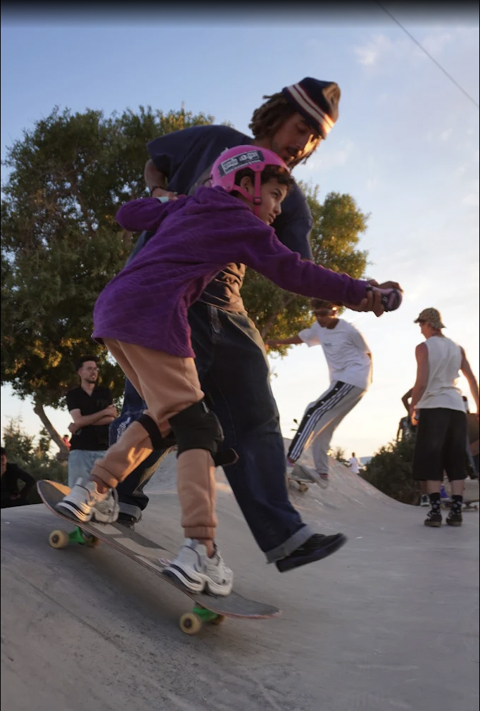 A coach helping a young skater balance on a skateboard at a concrete skatepark in Casablanca